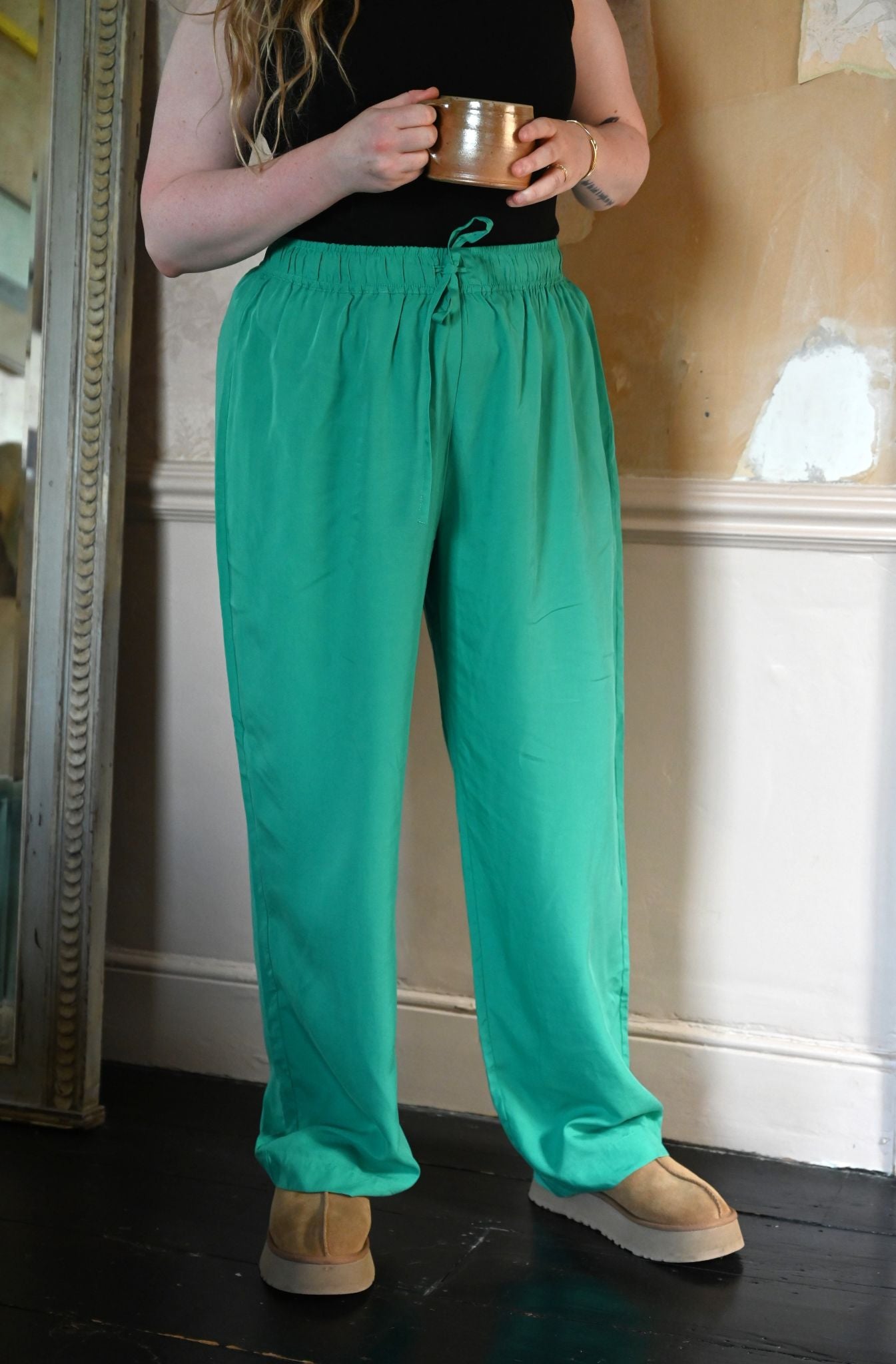 Person wearing teal pants and beige shoes holding a metallic cup indoors.