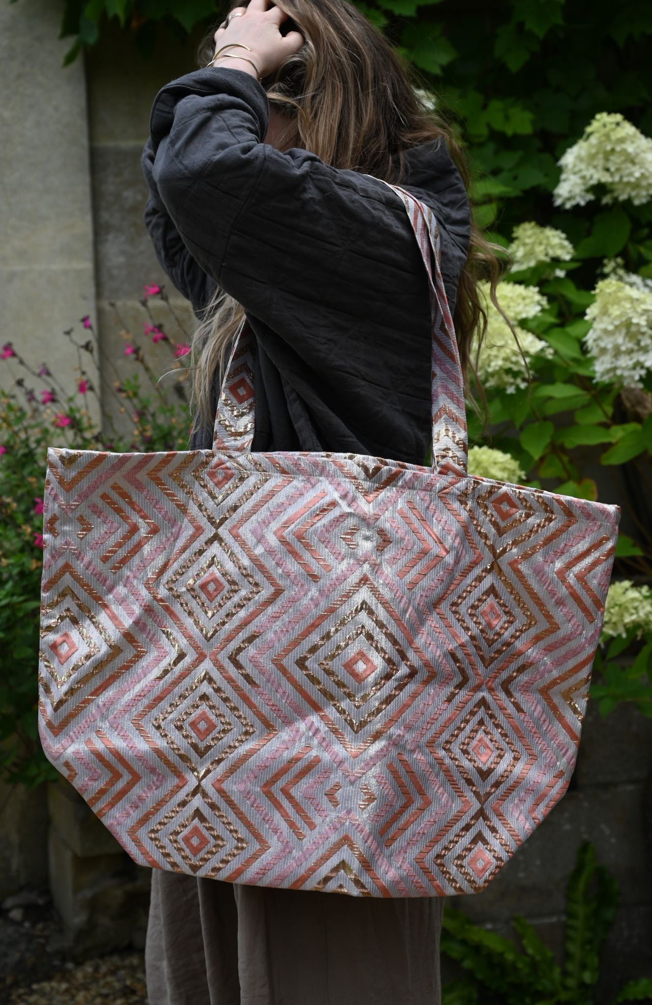Person holding a patterned tote bag outdoors with greenery and flowers in the background
