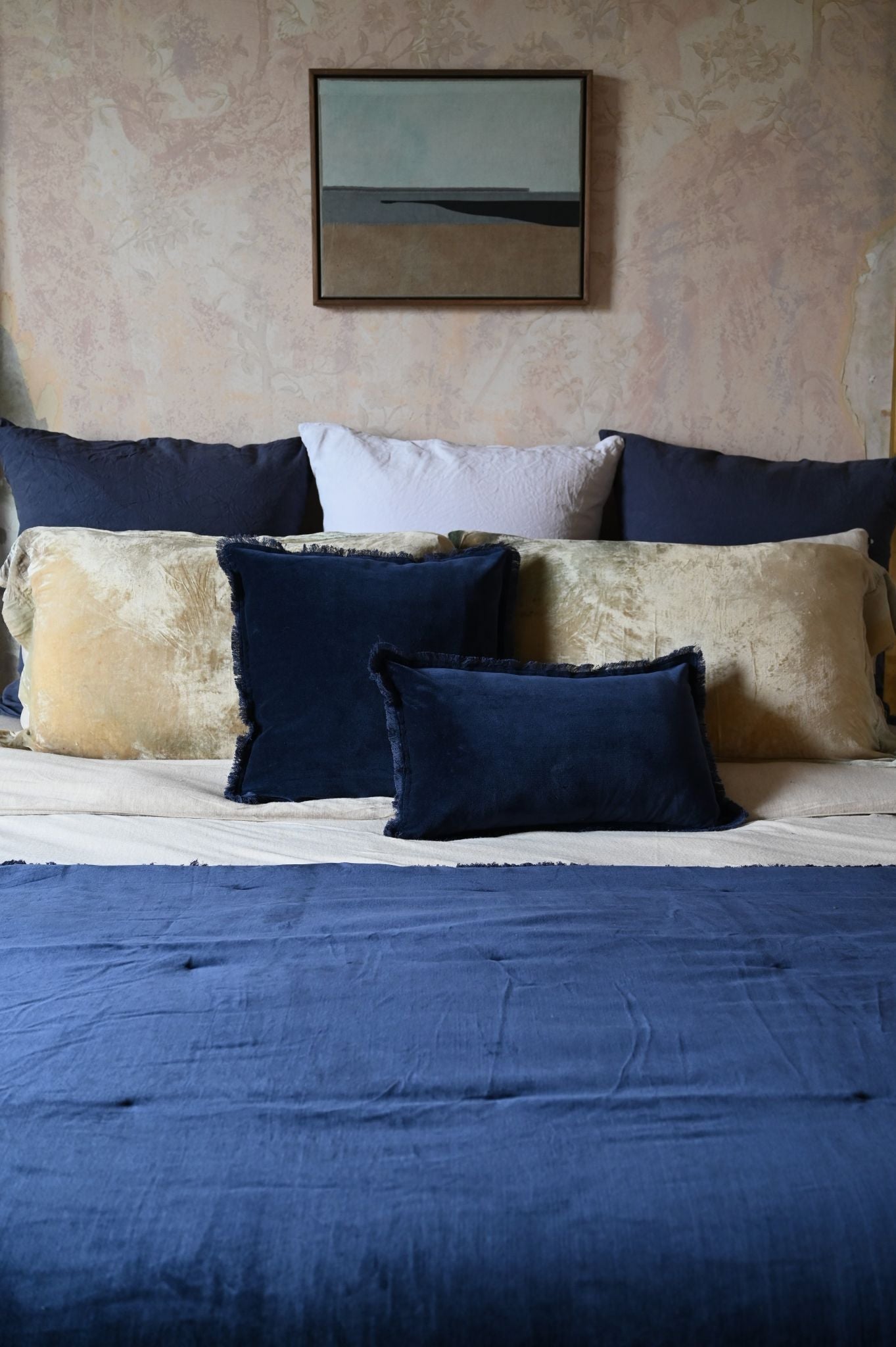 Deep navy fringed velvet cushions layered on a bed with gold and linen pillows.