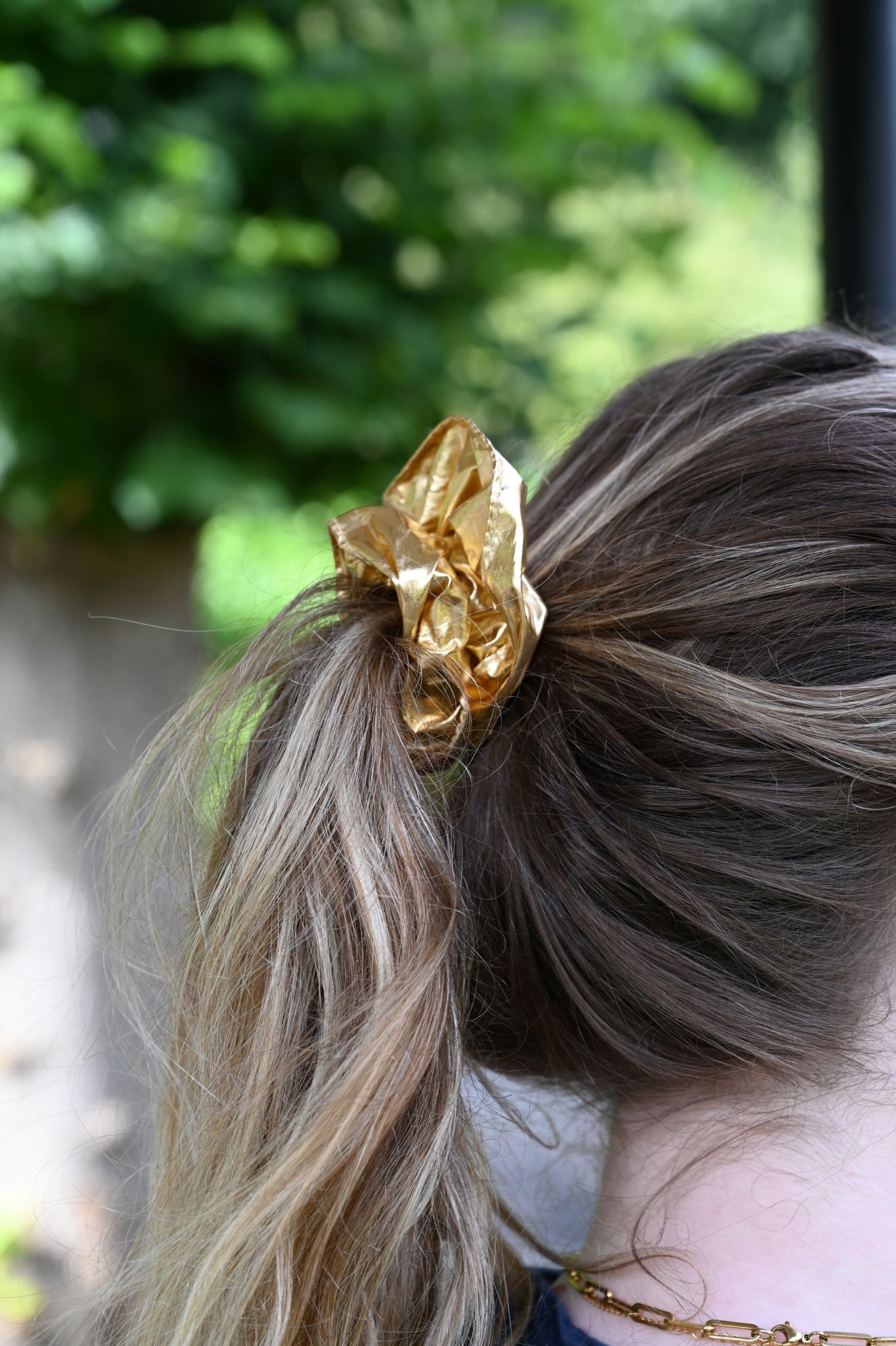 Chunky metallic gold scrunchie worn in a high ponytail against green background.