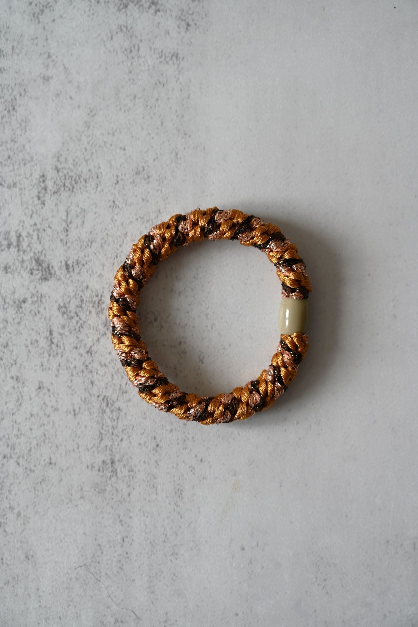 Hair tie with braided design and a central bead on a textured gray surface