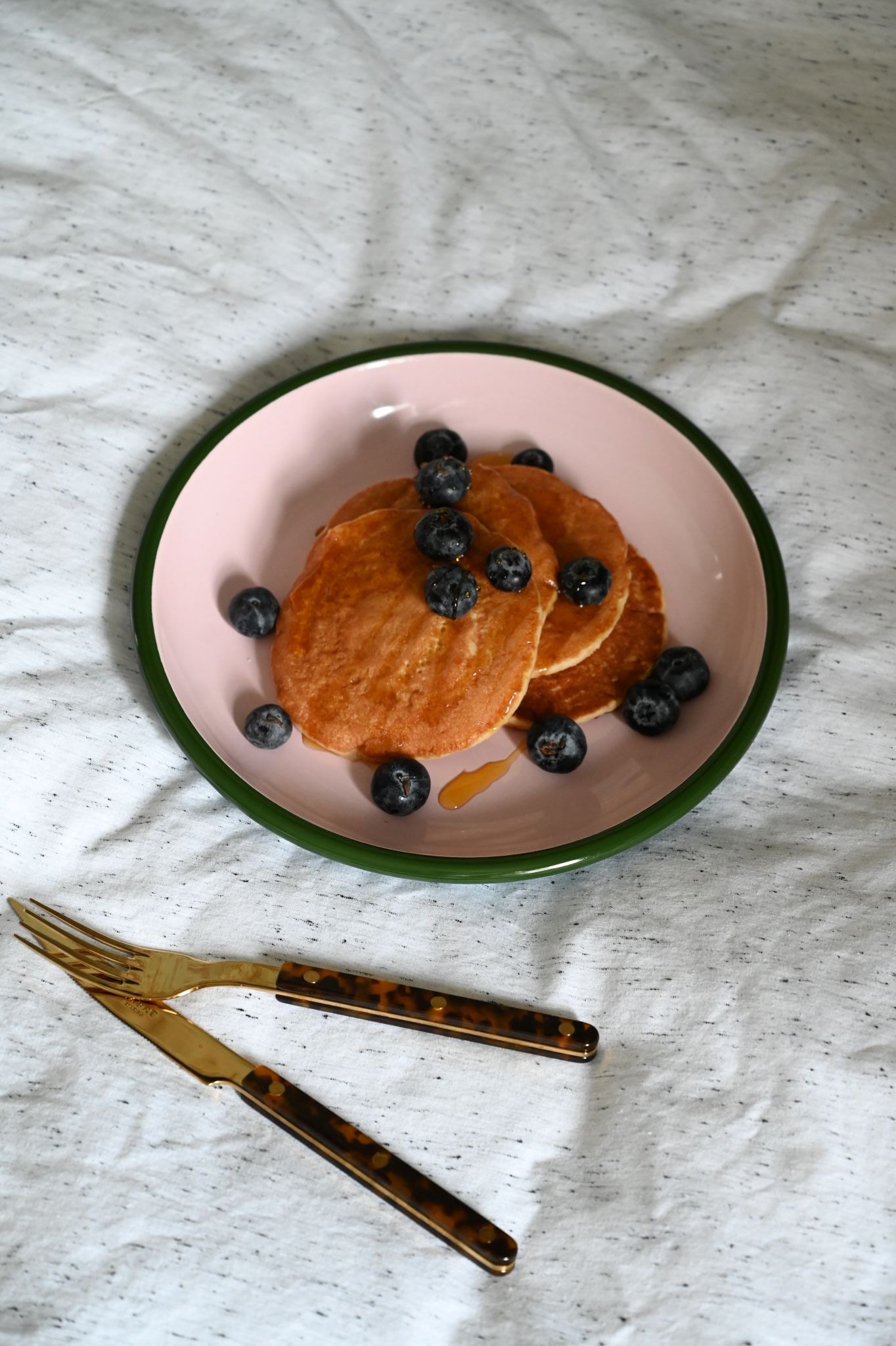 Stack of pancakes with blueberries on a pink pasta plate with a green rim, on a white textured surface.