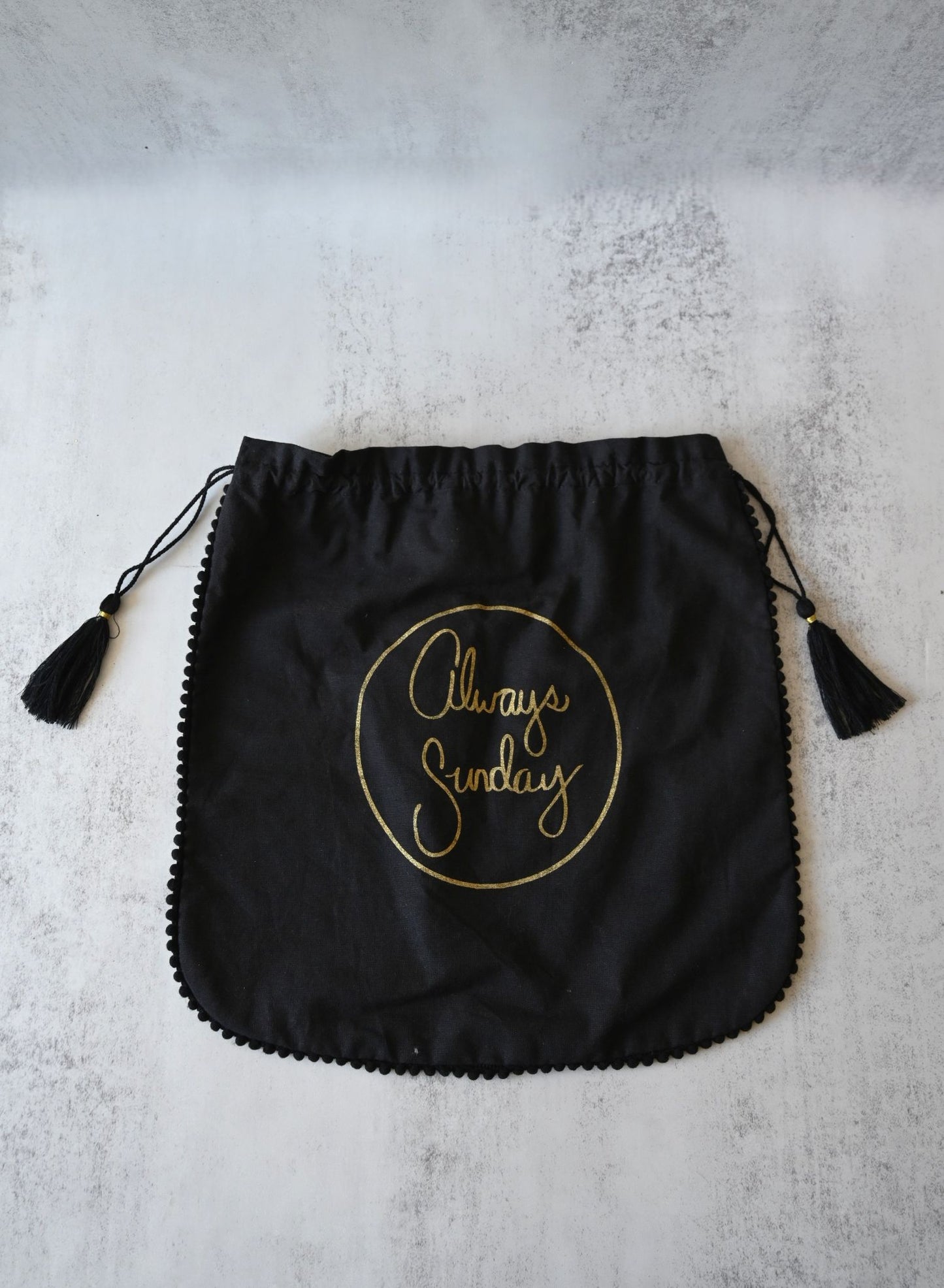 Large black cotton drawstring pouch with metallic gold Always Sunday script logo and tassel trim.