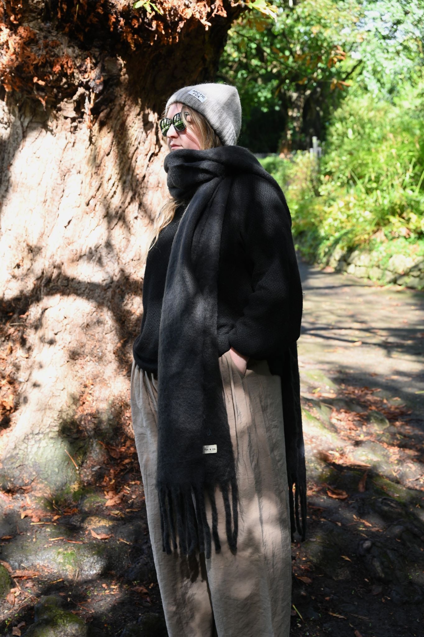 Model wearing oversized washed black recycled scarf with fringe in woodland.