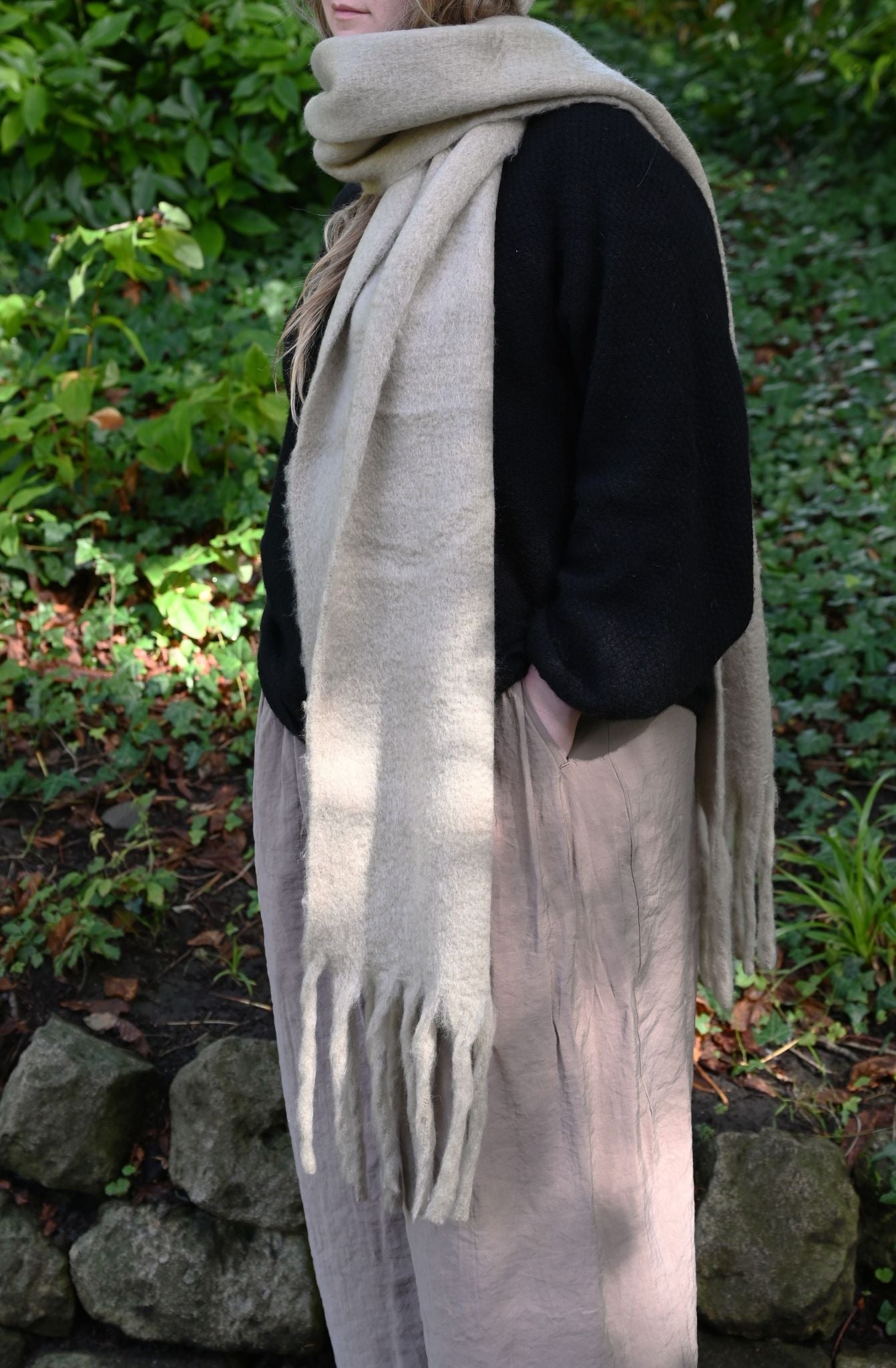 Oversized pebble beige fringed scarf worn loosely over a black jumper outdoors.