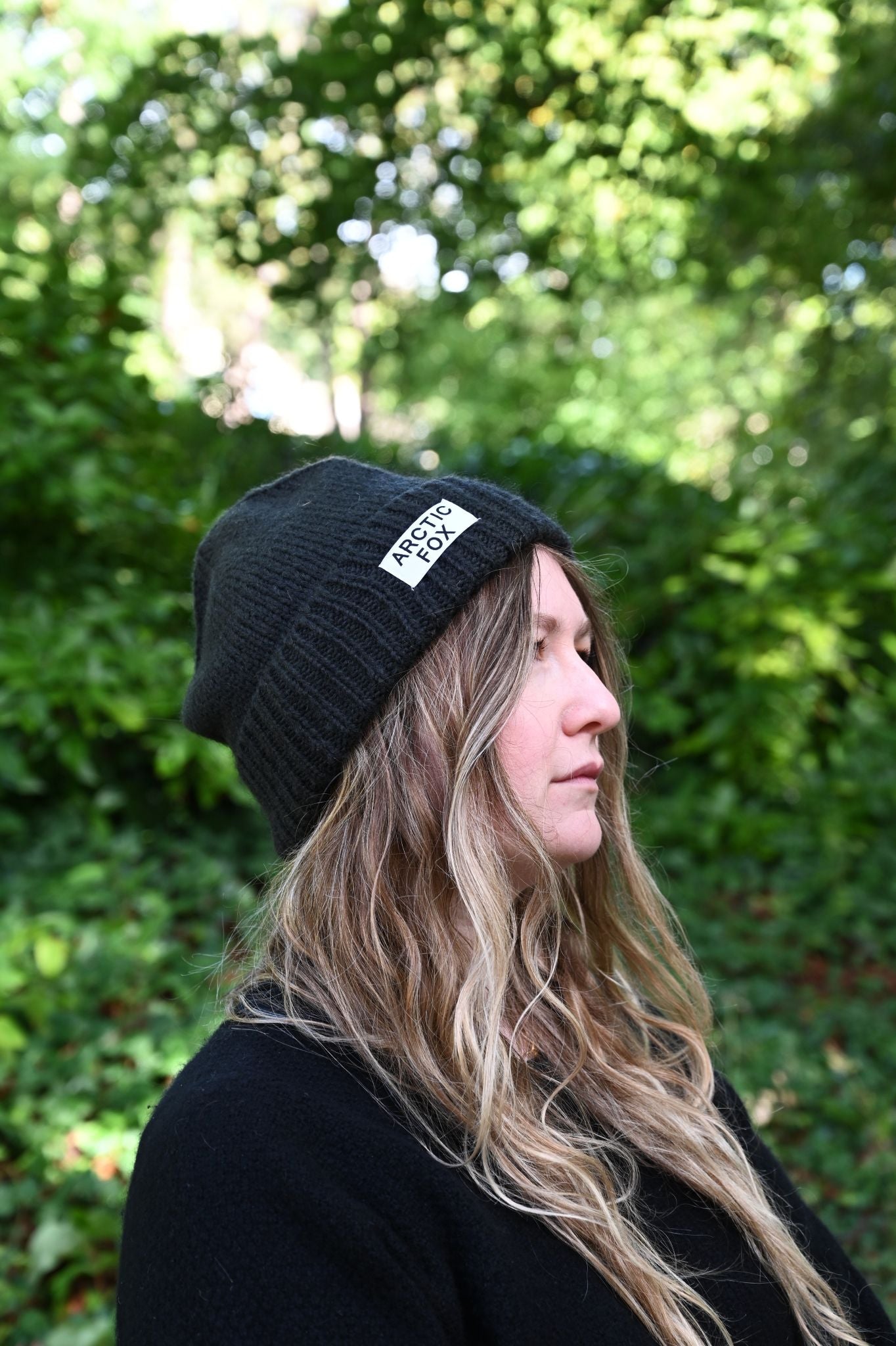 Person wearing a black beanie with a logo in front of green foliage