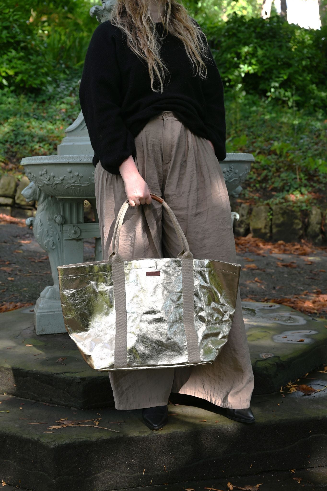 Person holding a shiny pale gold tote bag outdoors with greenery in the background