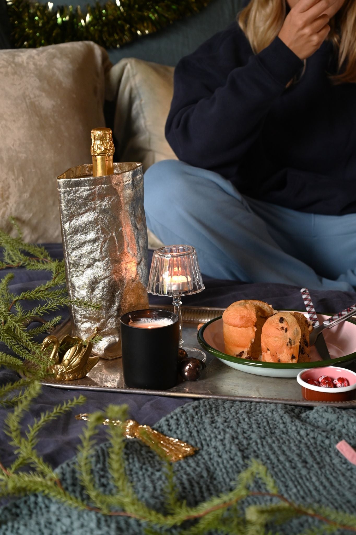 Person sitting on a bed with a tray of food and drinks, including a bottle of champagne. Enamel pasta plate, enamel sauce pot, uashmama wine cooler, clear glass lamp t-ligh and give us a snog candle on a mirrored tray.