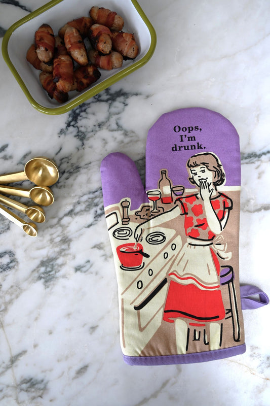 Oven mitt with a humorous design of a woman and  'Oops I'm drunk' text, placed on a marble surface with a container of sausages and measuring spoons.
