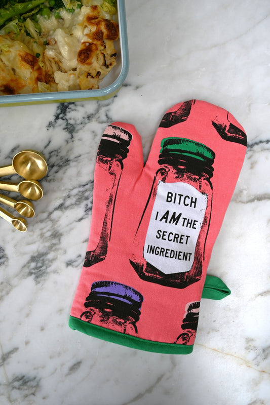 Oven mitt with graphic design and 'Bitch I am the secret ingredient' text on a marble surface with food items.