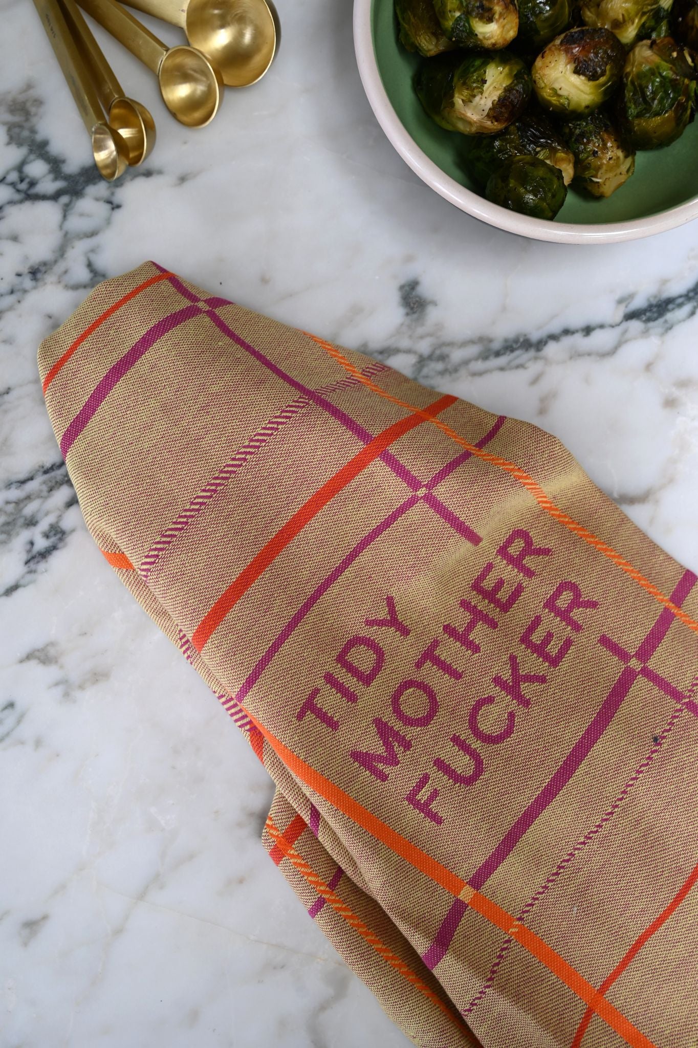 Beige tea towel with plaid pattern and tidy mother fucker text on a marble surface with brussels sprouts and gold measuring spoons.