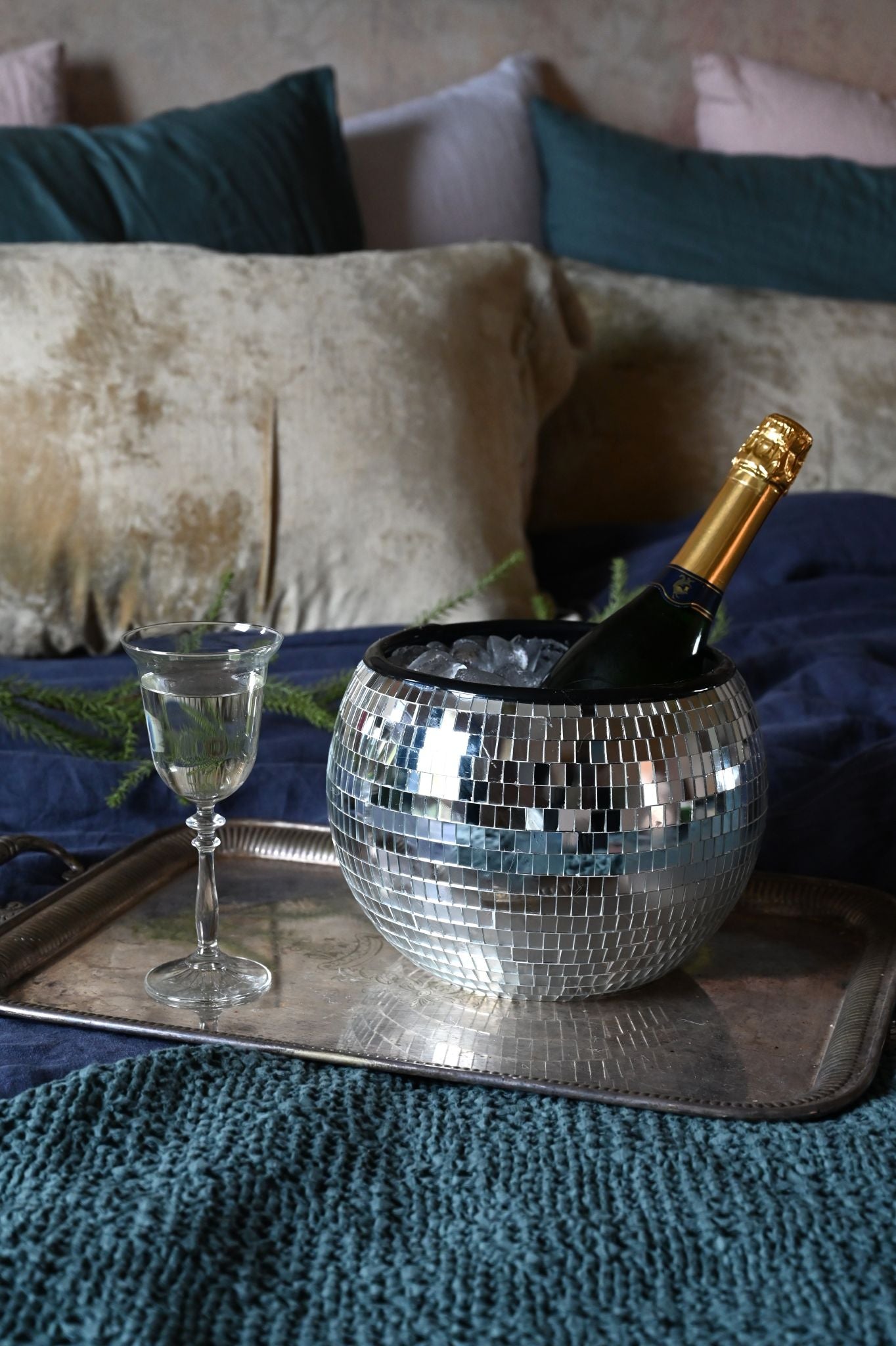 Champagne bottle in a disco ball on a tray with a glass, set against a textured surface with pillows.