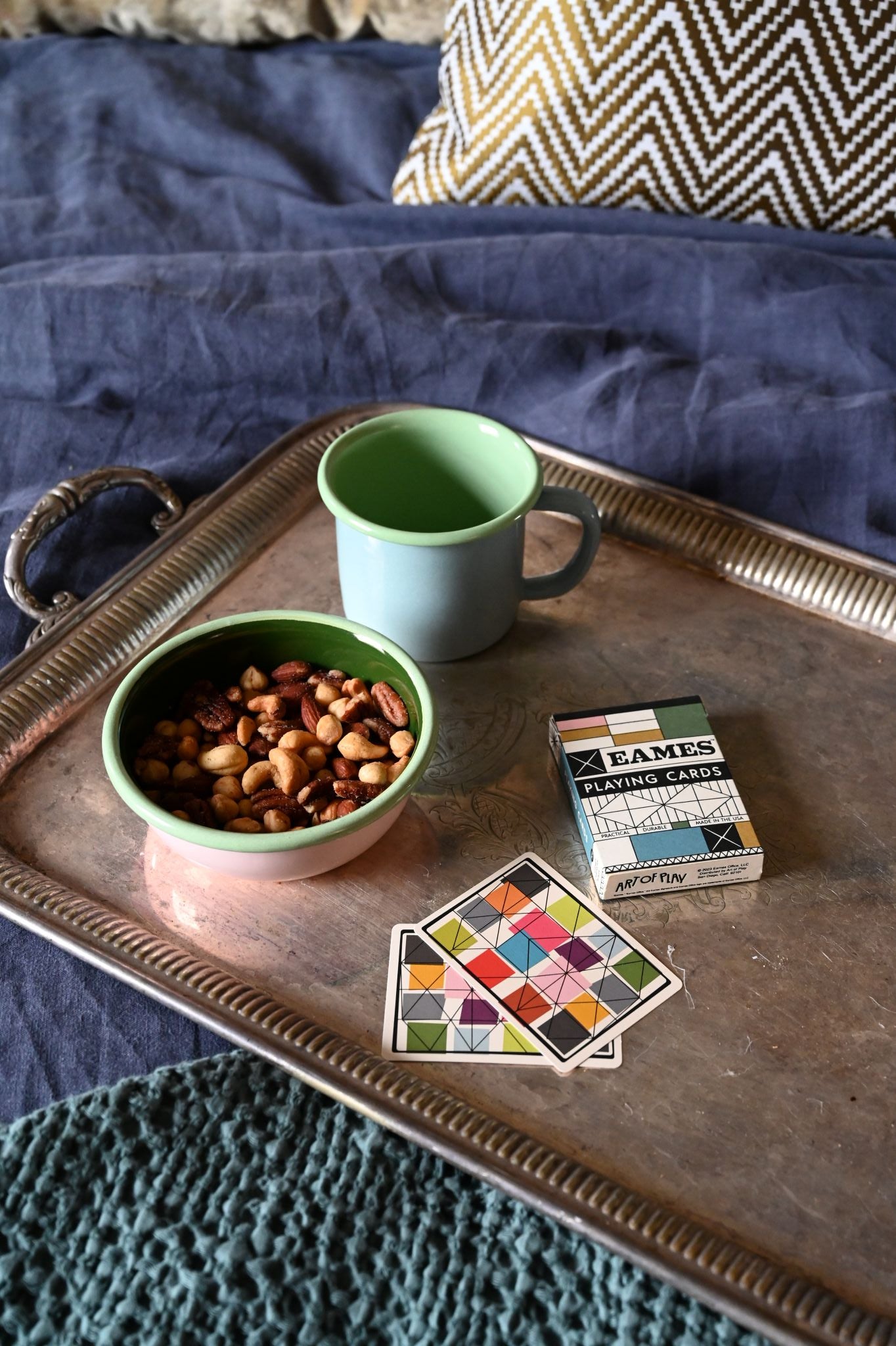 Large enamel mug and small enamel bowl with snacks on a metal tray, next to a box of playing cards on a textured surface.