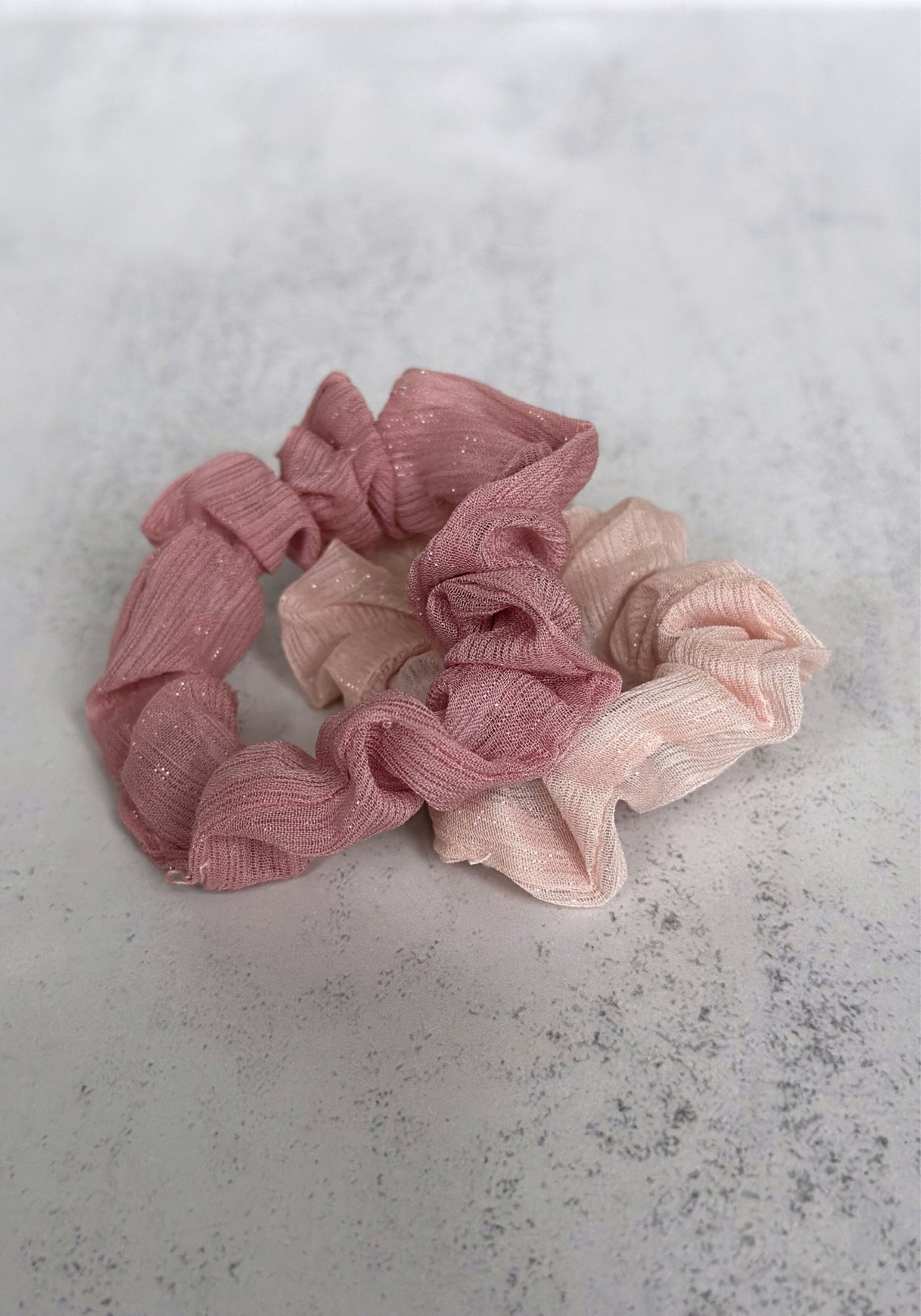 Two small pink scrunchies tied in a knot, placed on a textured grey surface.