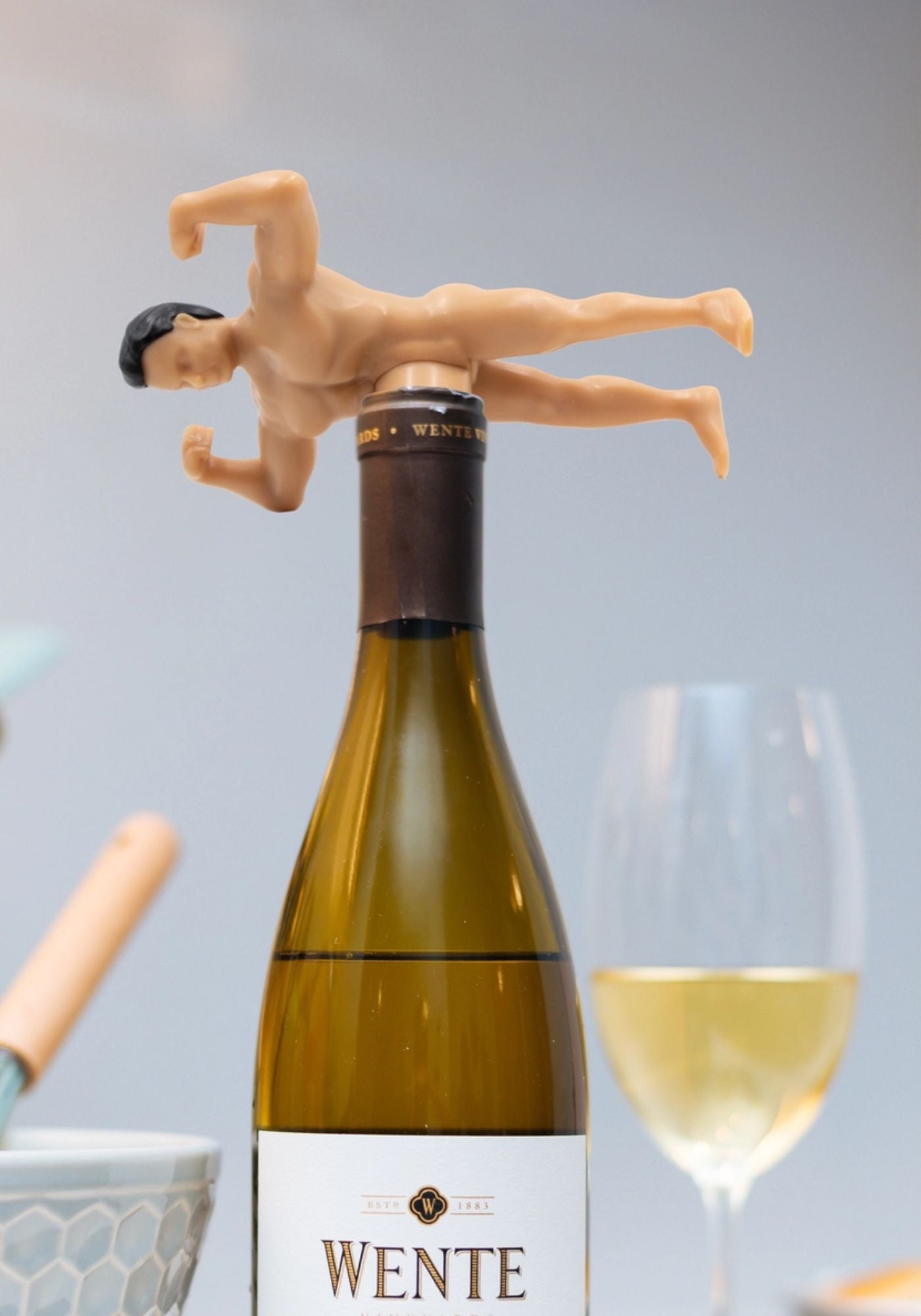 Wine bottle with a humorous figurine on top, next to a glass of white wine.