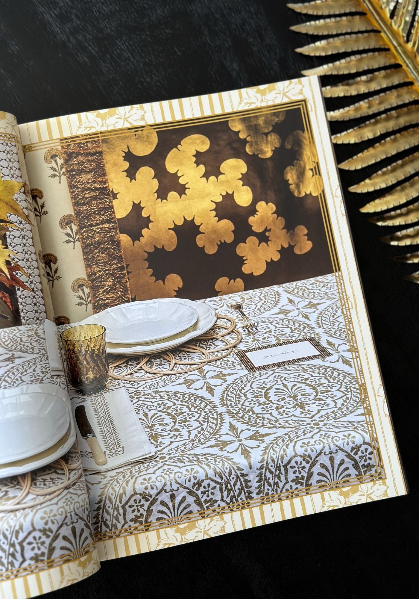 Decorative table setting with gold and black patterned tablecloth, plates, and cutlery on a black background.