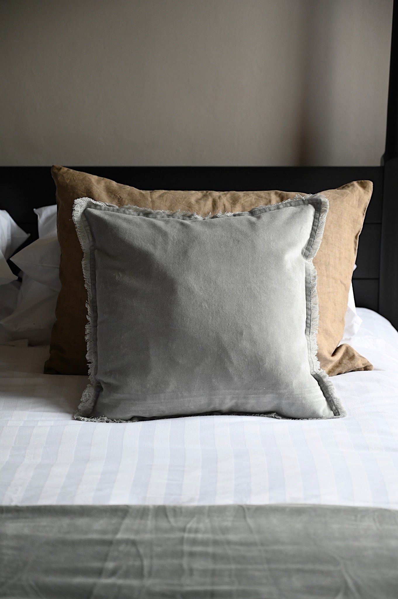 Layered pearl grey fringed velvet cushion on a bed with neutral linens.