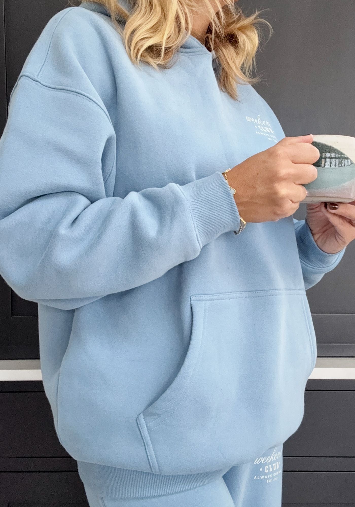 Person wearing a light blue hoodie holding a white mug with a dark interior.