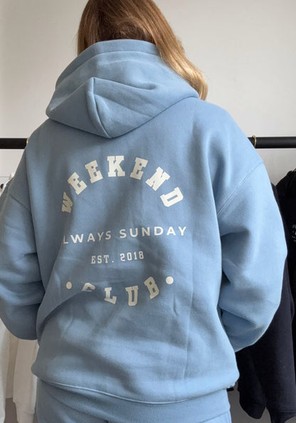 Light blue hoodie with text on the back worn by a person, against a neutral background.