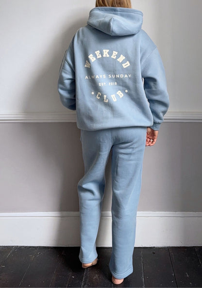 Person wearing a light blue hoodie and pants with text on the back against a gray wall.