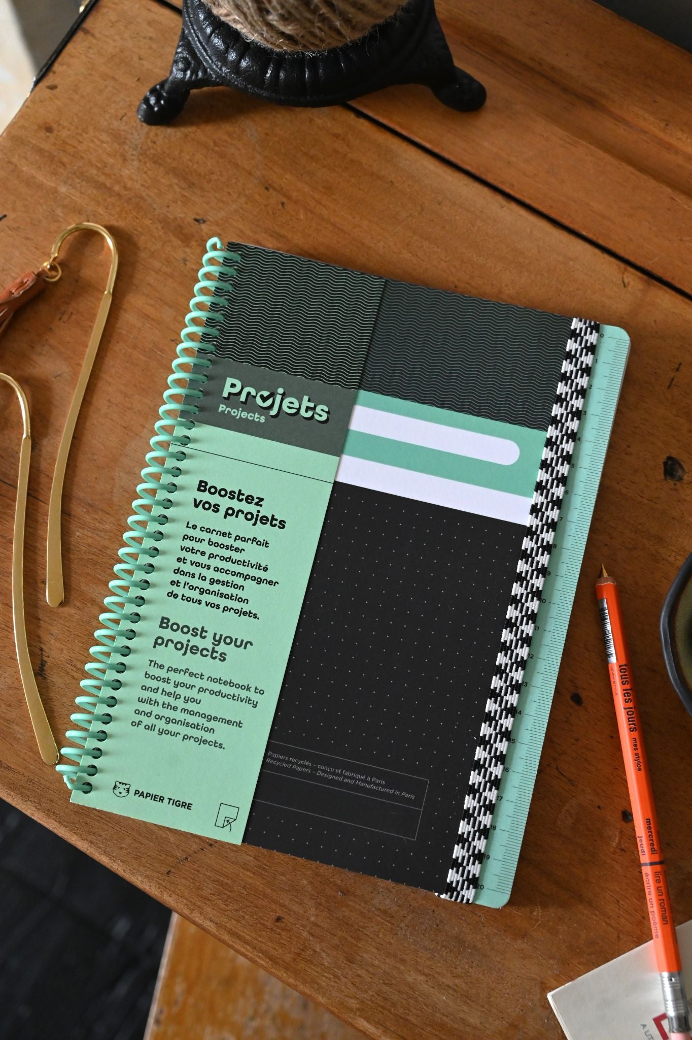 A5 Project Planner Notebook lying on a wooden surface with a pen and decorative items in the periphery. The notebook has a black and green cover with white spiral binding.