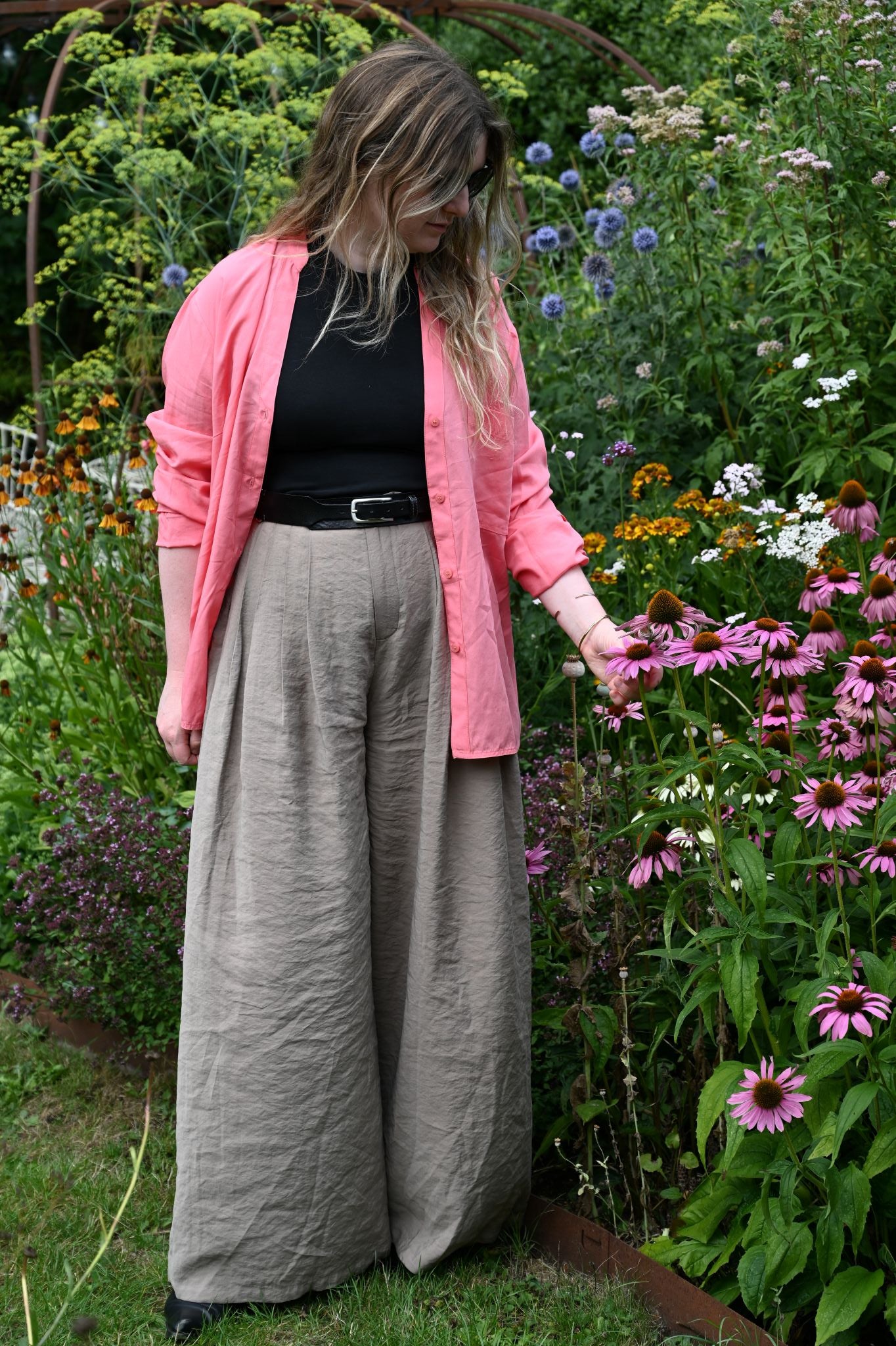 Person wearing gray pants and a pink cardigan standing in a garden with flowers.