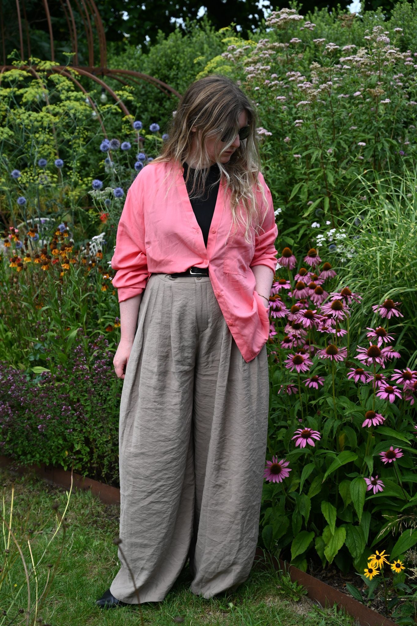 Person wearing a pink jacket and beige pants standing in a garden with flowers and greenery.
