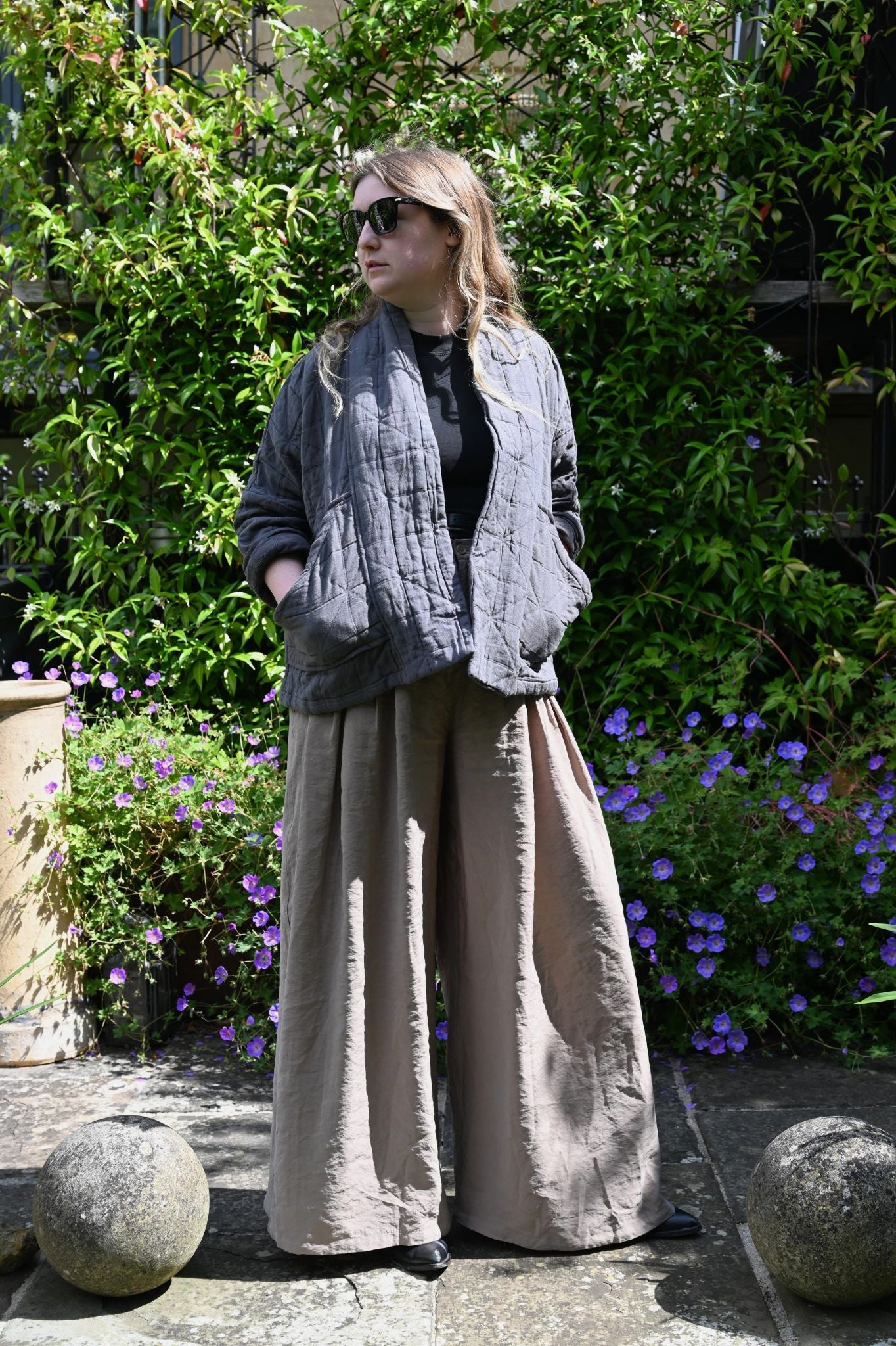 Person wearing a gray quilted jacket and beige pants standing in front of greenery with purple flowers.