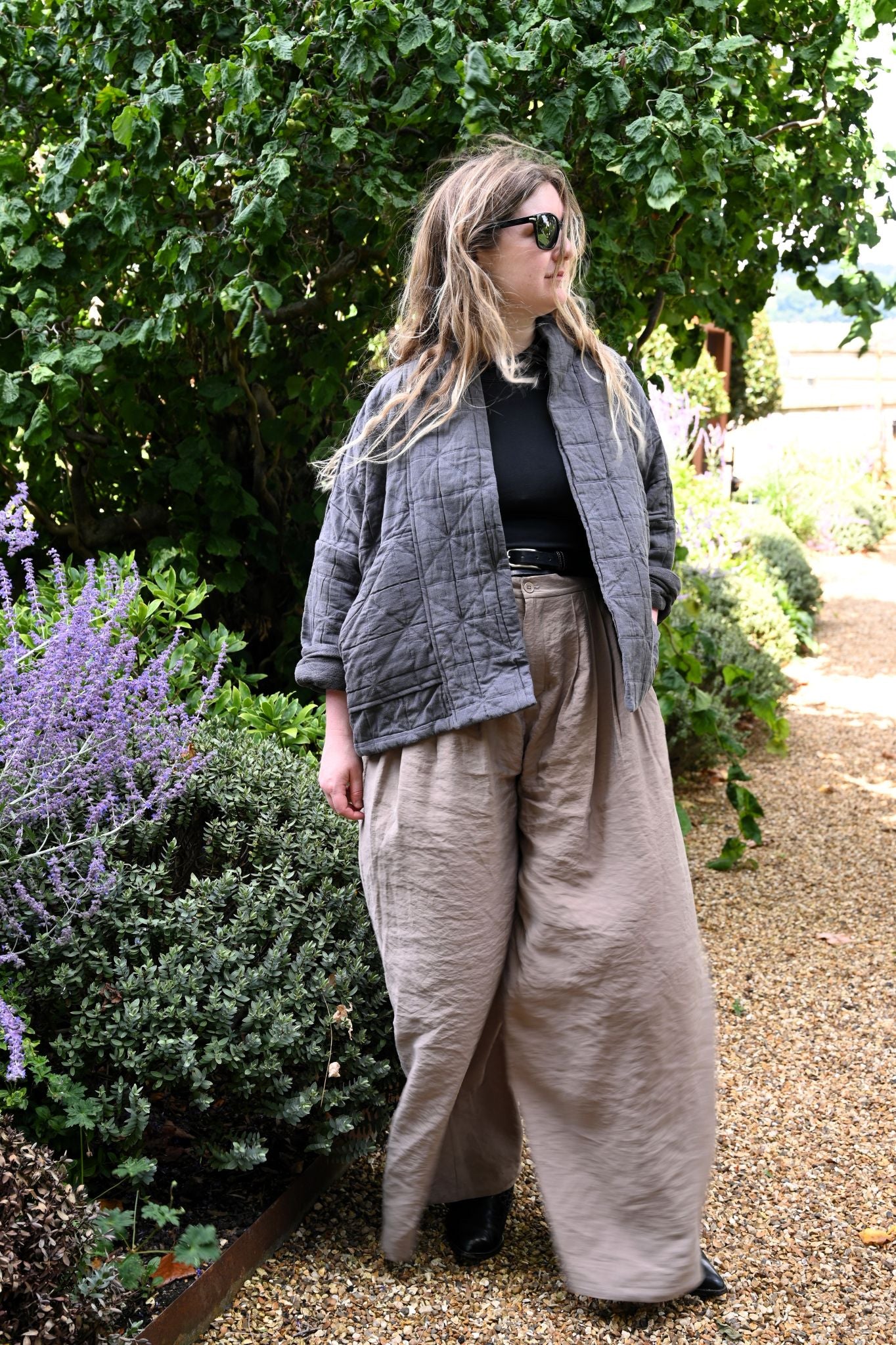 Person wearing a gray jacket and beige pants standing in a garden with greenery and flowers.