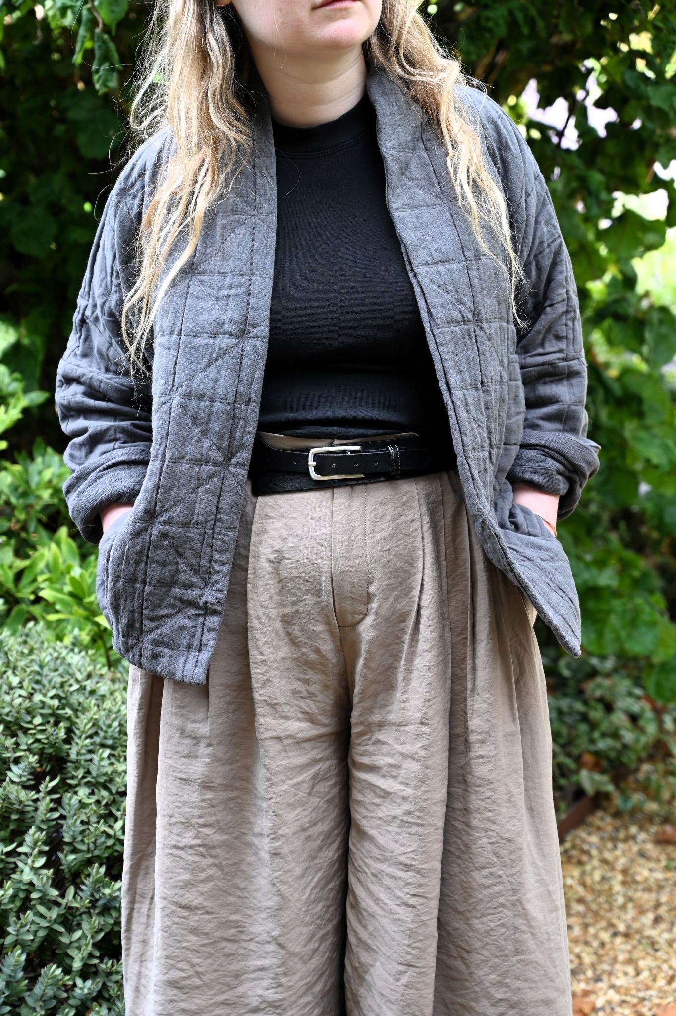 Person wearing a gray quilted jacket, black top, and beige pants with a green outdoor background