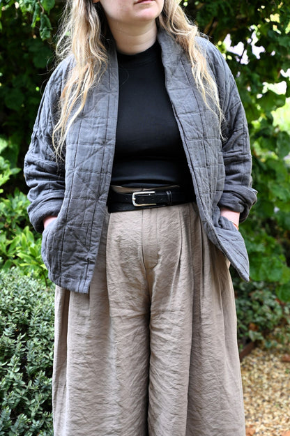 Person wearing a gray quilted jacket, black top, and beige pants with a green outdoor background