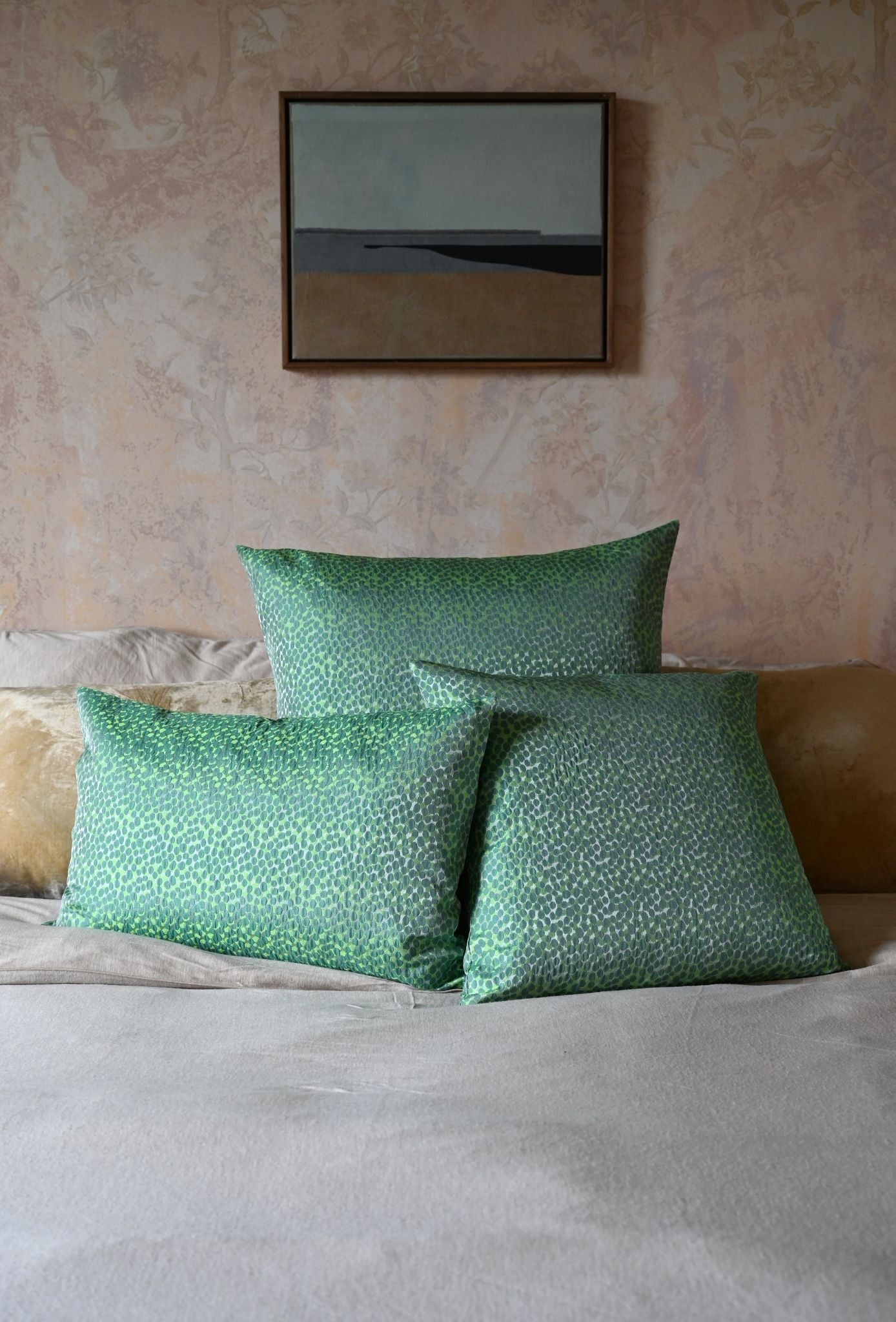Green patterned pillows on a bed with a neutral wall and framed picture in the background