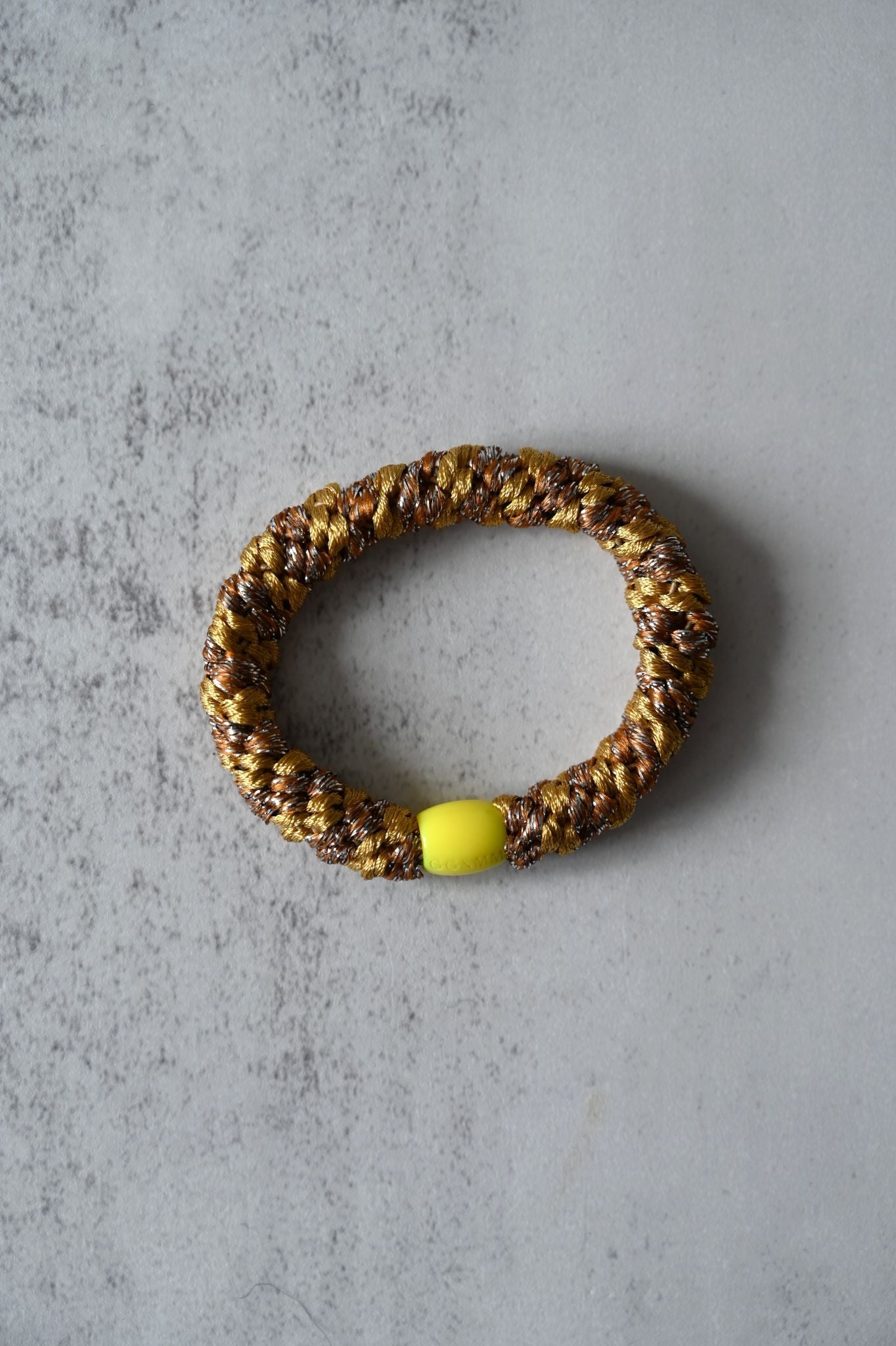Hair tie with gold and brown pattern and a yellow bead on a gray surface