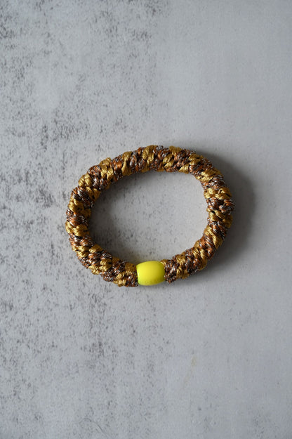 Hair tie with gold and brown pattern and a yellow bead on a gray surface