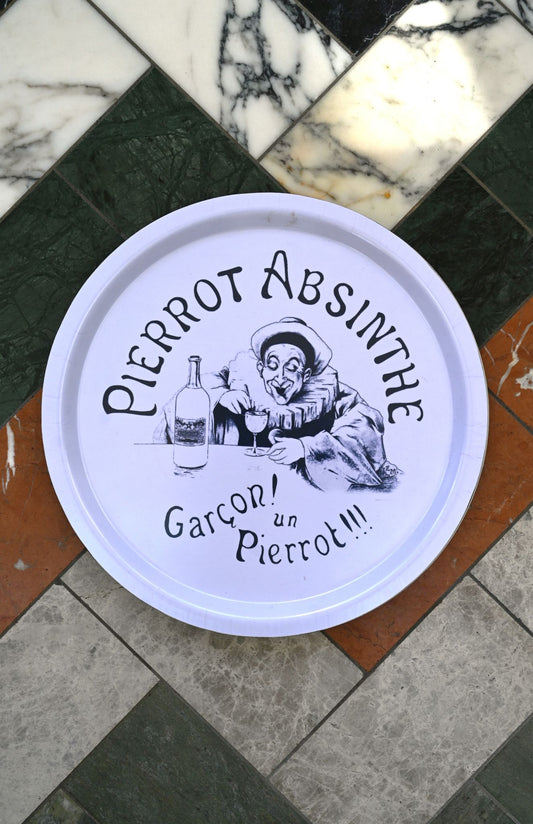 Round white sign with black text and illustration of a Pierrot Absinthe character on a marble floor.