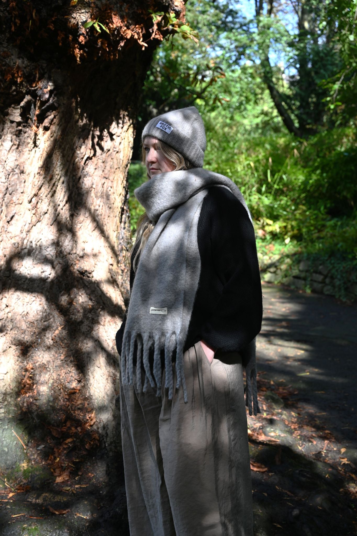 Person wearing a gray scarf and hat standing next to a tree in a forest