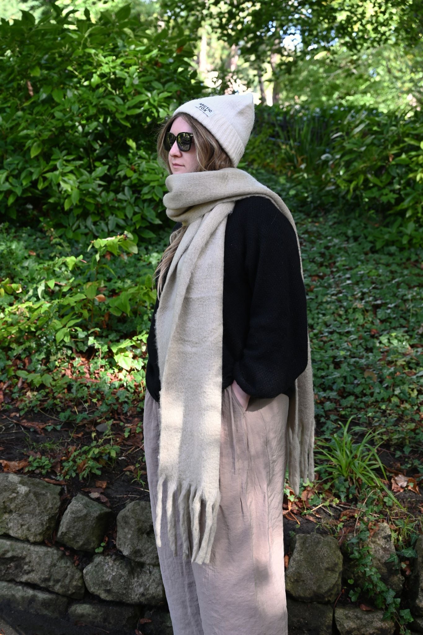 Person wearing a beige scarf, black jacket, and white beanie in a forest setting.