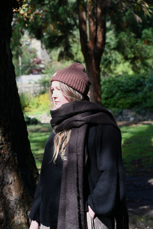 Person wearing a brown beanie and dark brown scarf standing in a park-like setting with trees and grass.