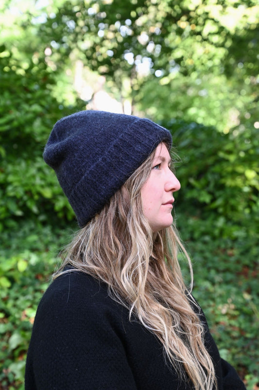 Person wearing a dark blue knit beanie and coat outdoors with greenery in the background