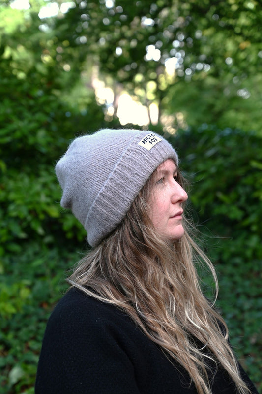 Person wearing a gray beanie with a logo, standing in front of green foliage.