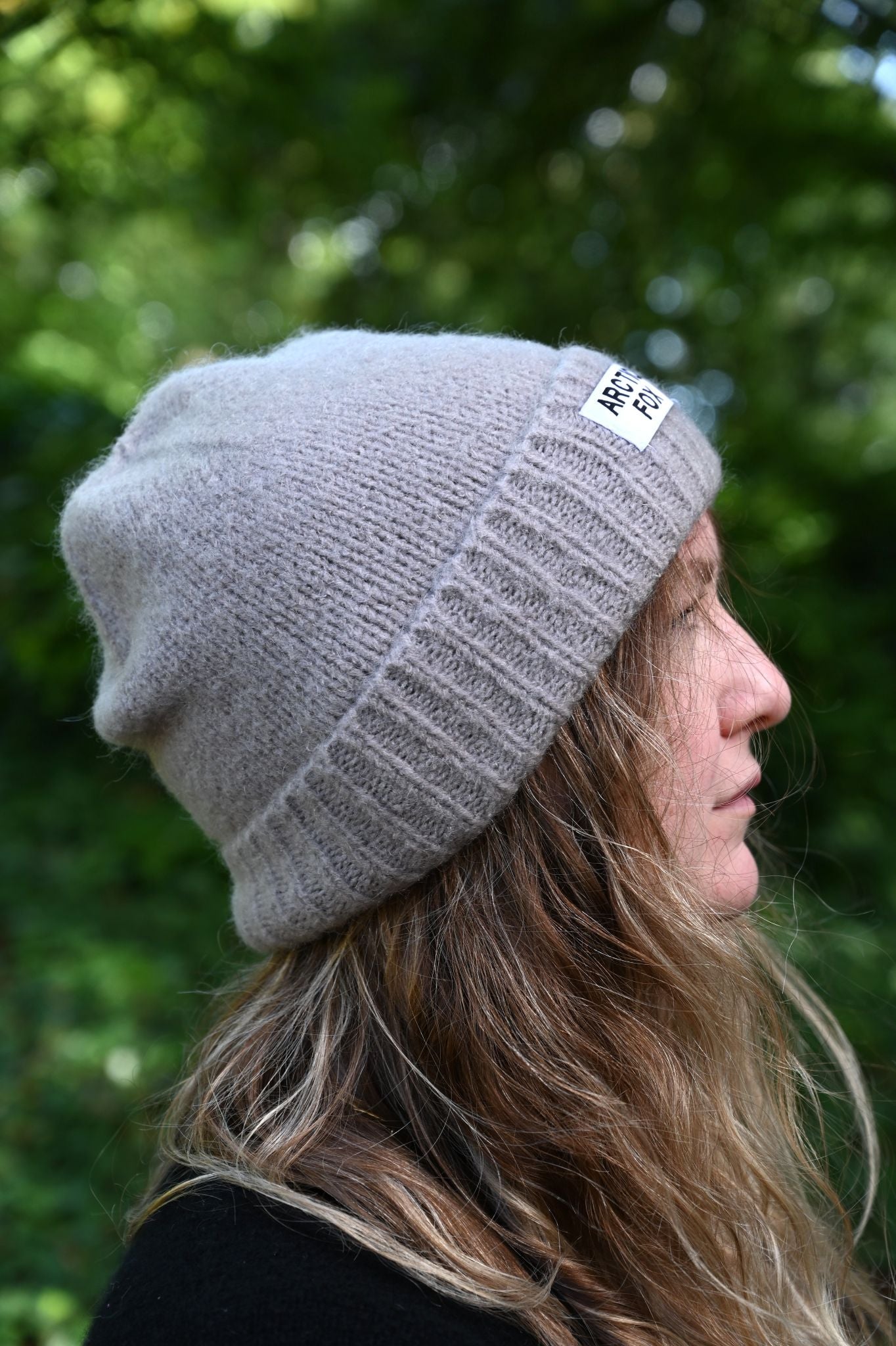 Person wearing a gray knit beanie with a logo, standing outdoors with greenery in the background