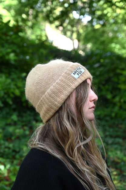 Person wearing a beige beanie with a logo, standing outdoors against a green leafy background