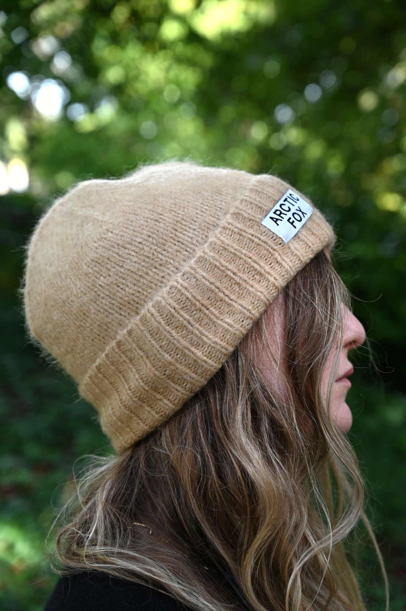 Beige knit beanie with a brand label worn by a person outdoors.