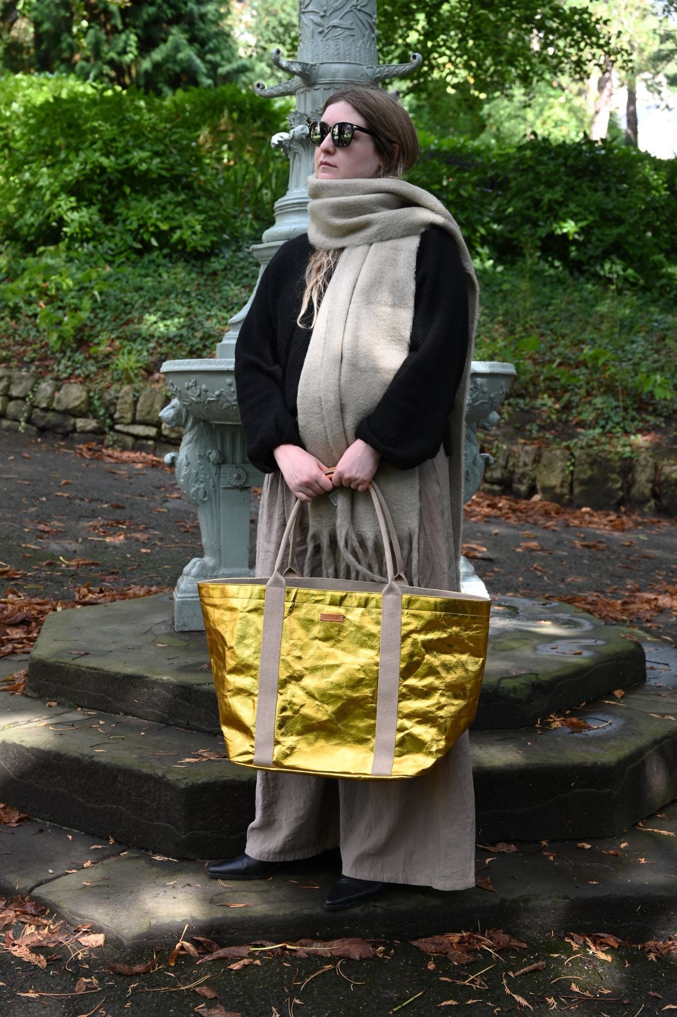 Person holding a gold tote bag in an outdoor setting with greenery