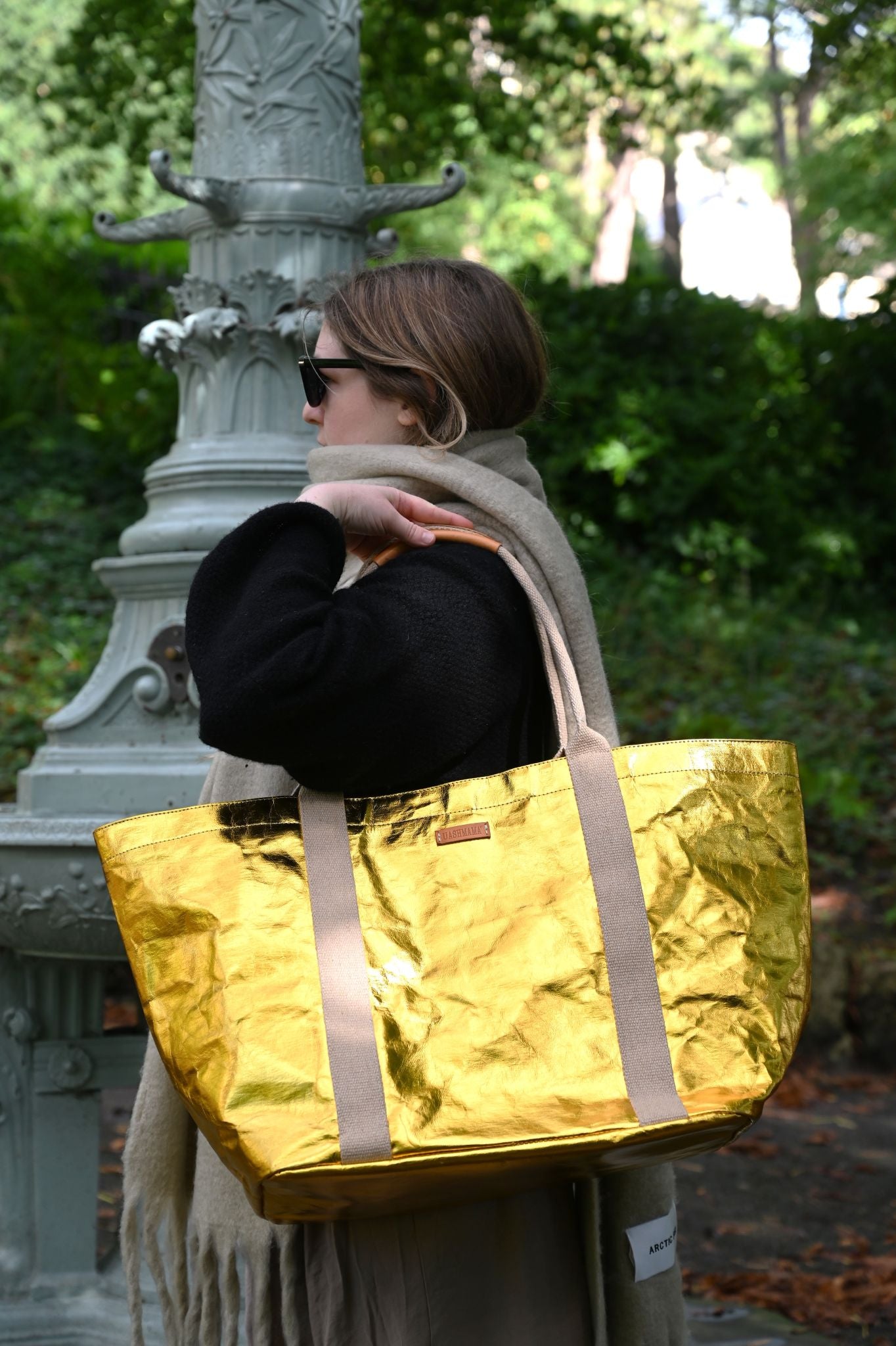 Person holding a gold tote bag outdoors with greenery in the background