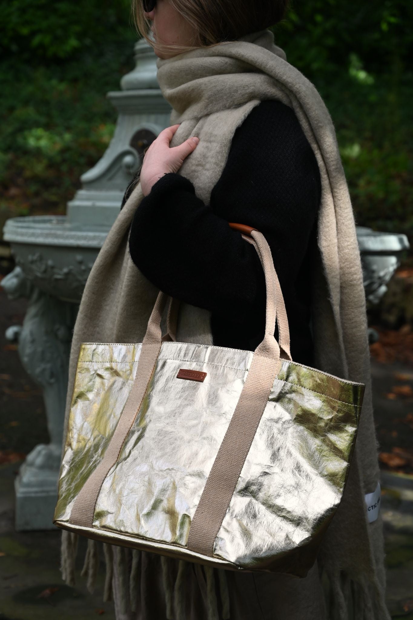 Person holding a small pale gold tote bag, wearing a scarf outdoors.