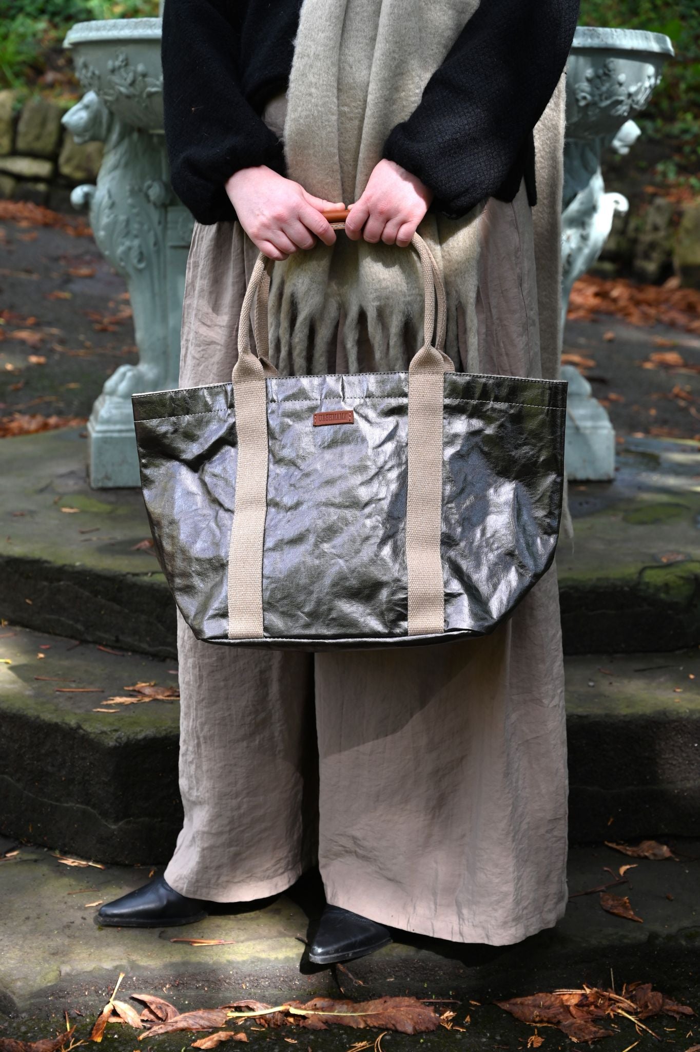 Person holding a small gunmetal tote bag outdoors on a stone path.