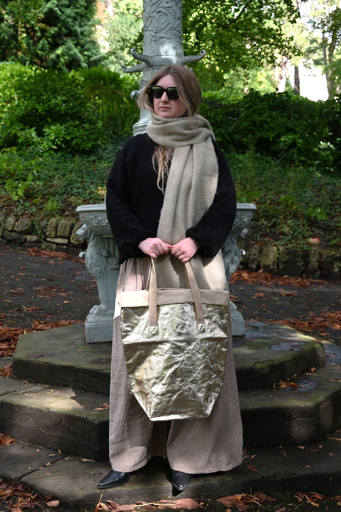 Person holding a large metallic pale gold bag in an outdoor setting with greenery.