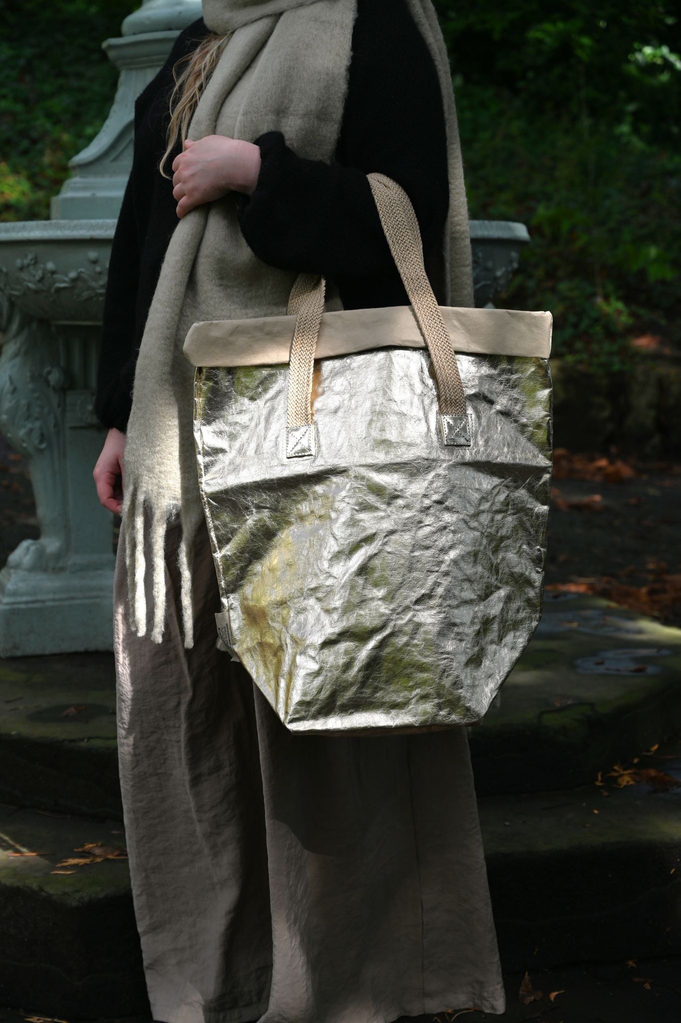 Person holding a metallic pale gold bag outdoors with greenery in the background