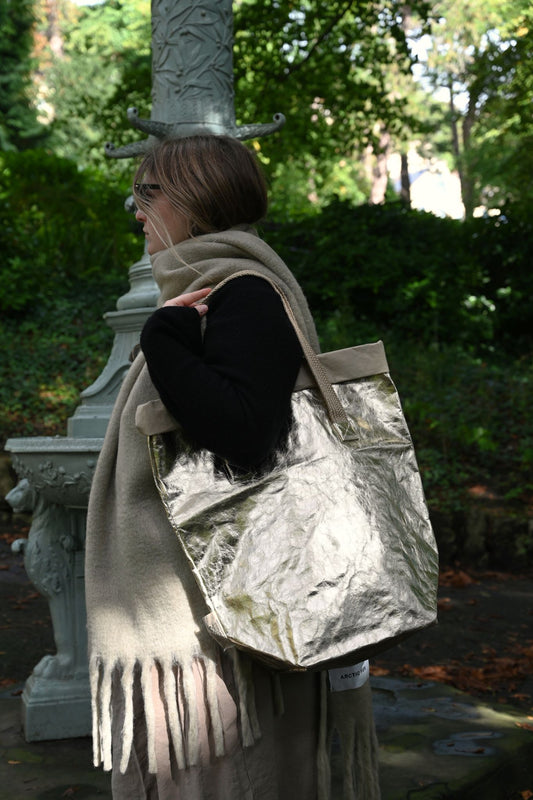 Person holding a pale gold metallic bag in an outdoor setting with greenery.